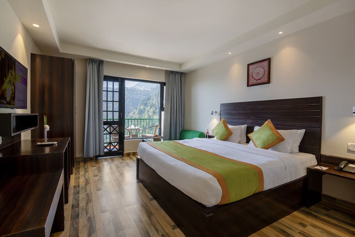 A hotel room with a large bed, a desk, a chair, a balcony with a view of the mountains, and a framed picture on the wall.