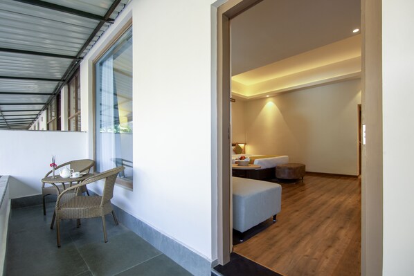 Una habitación de hotel moderna con balcón, una zona de comedor con sillas de mimbre, una cama con mesita de noche y suelo de madera.