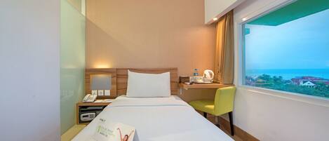 A hotel room with a bed, a desk, a chair, a nightstand, a phone, a kettle, and a window with a view of buildings.