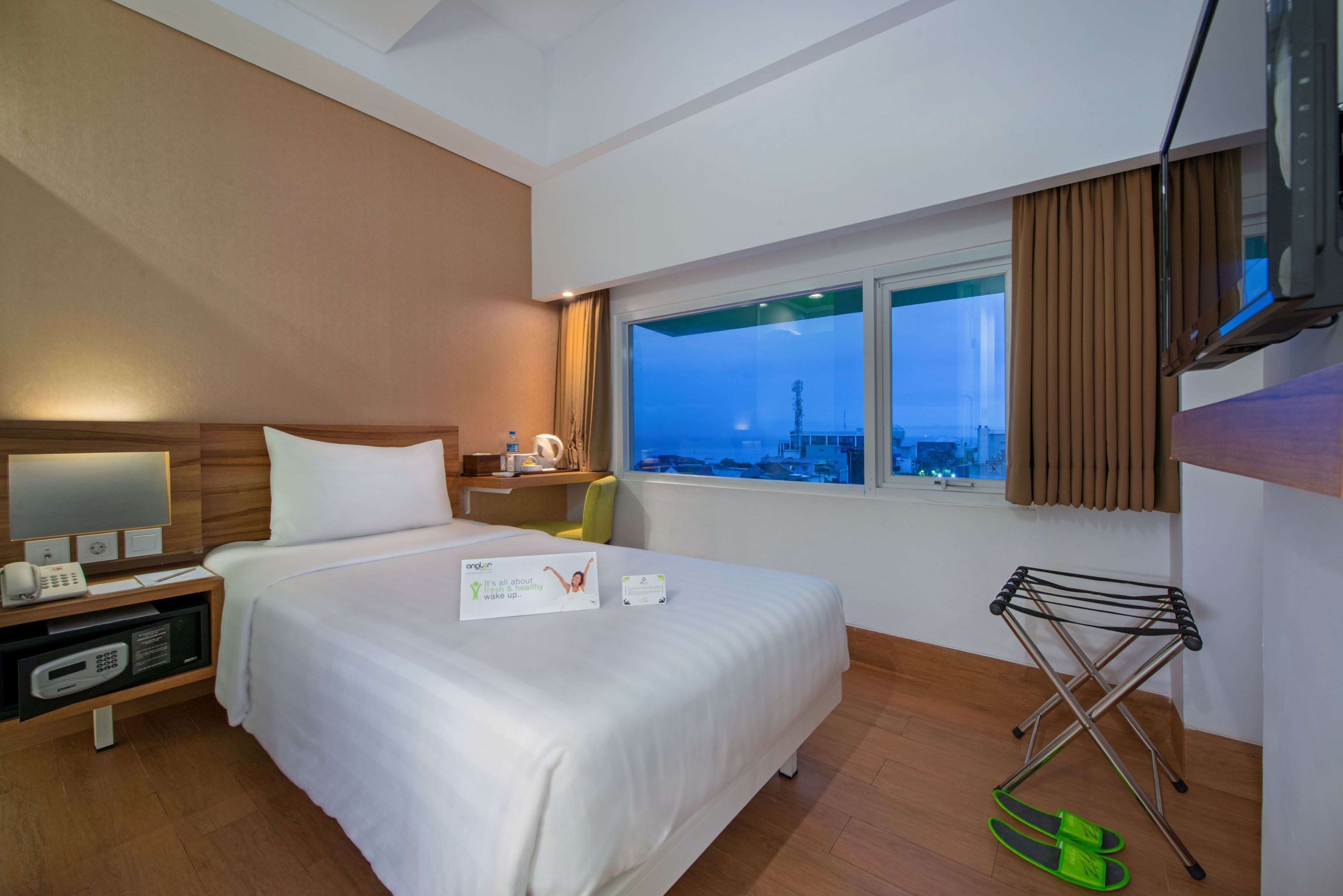 A hotel room with a bed, a nightstand with a phone and a digital device, a window with a view of a cityscape, and a drying rack with slippers.