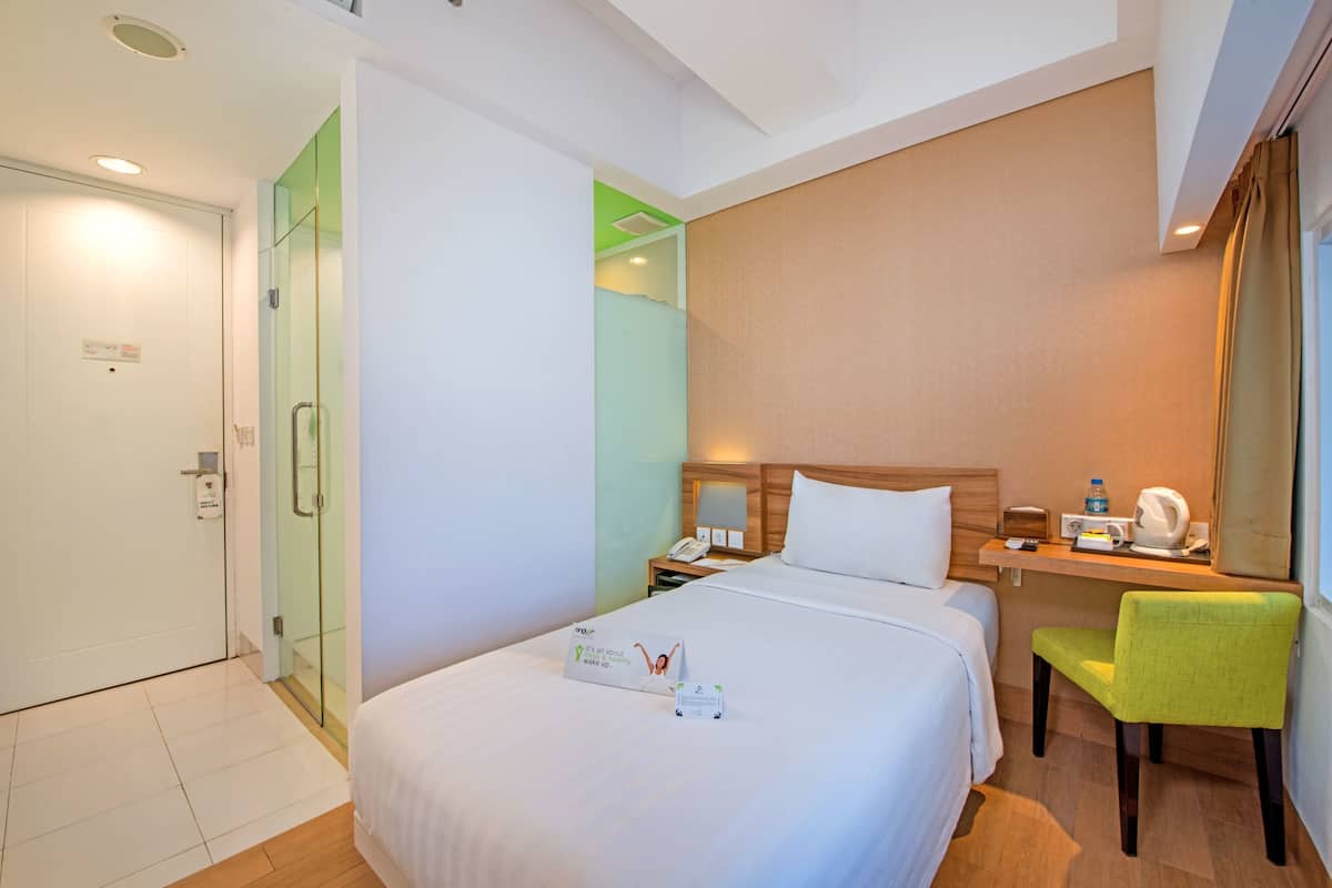 A hotel room with a bed, a desk with a chair, a nightstand, a green chair, a bedside table with a lamp, and a door with a glass panel.
