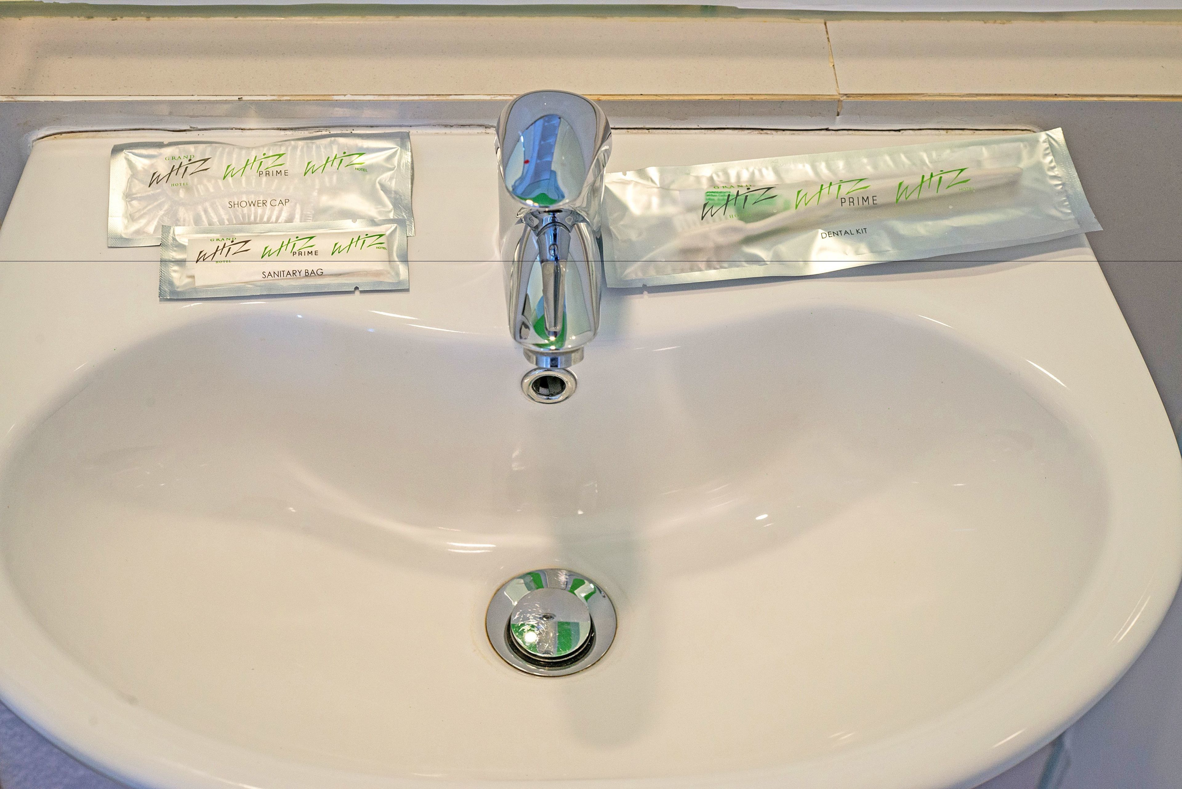 A white bathroom sink with a chrome faucet and a drain stopper. There are three items on the sink: a shower cap, a dental kit, and a sanitary bag.