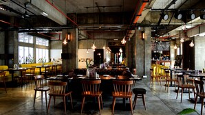 Un restaurante espacioso con techos altos, conductos expuestos y lámparas colgantes. Las mesas están dispuestas alrededor de una barra central.