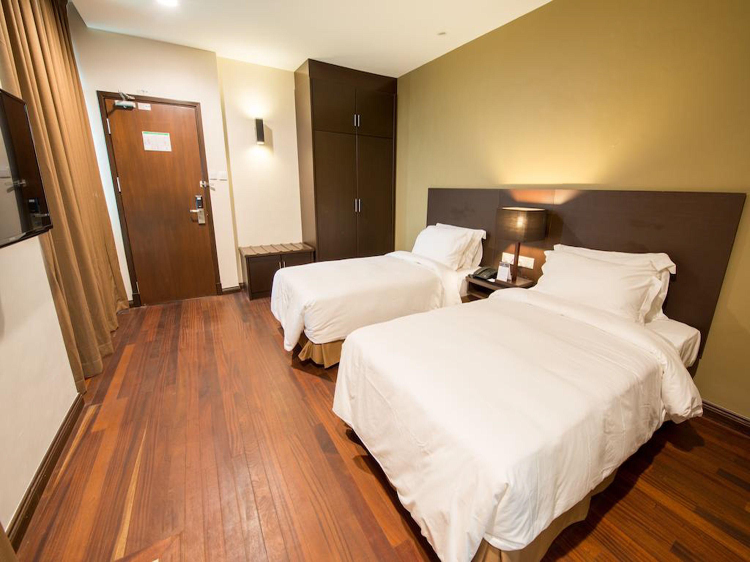 A hotel room with two beds, a wooden floor, a wardrobe, a desk with a lamp, and a door.