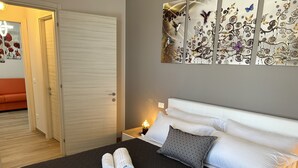 A hotel room with a bed, two towels, a nightstand, a wall art piece, and a door to another room. - Albergo Panoramica (Bardolino)
