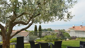 An outdoor seating area with wicker chairs and a table, overlooking a body of water and a distant view of buildings. - Albergo Panoramica (Bardolino)