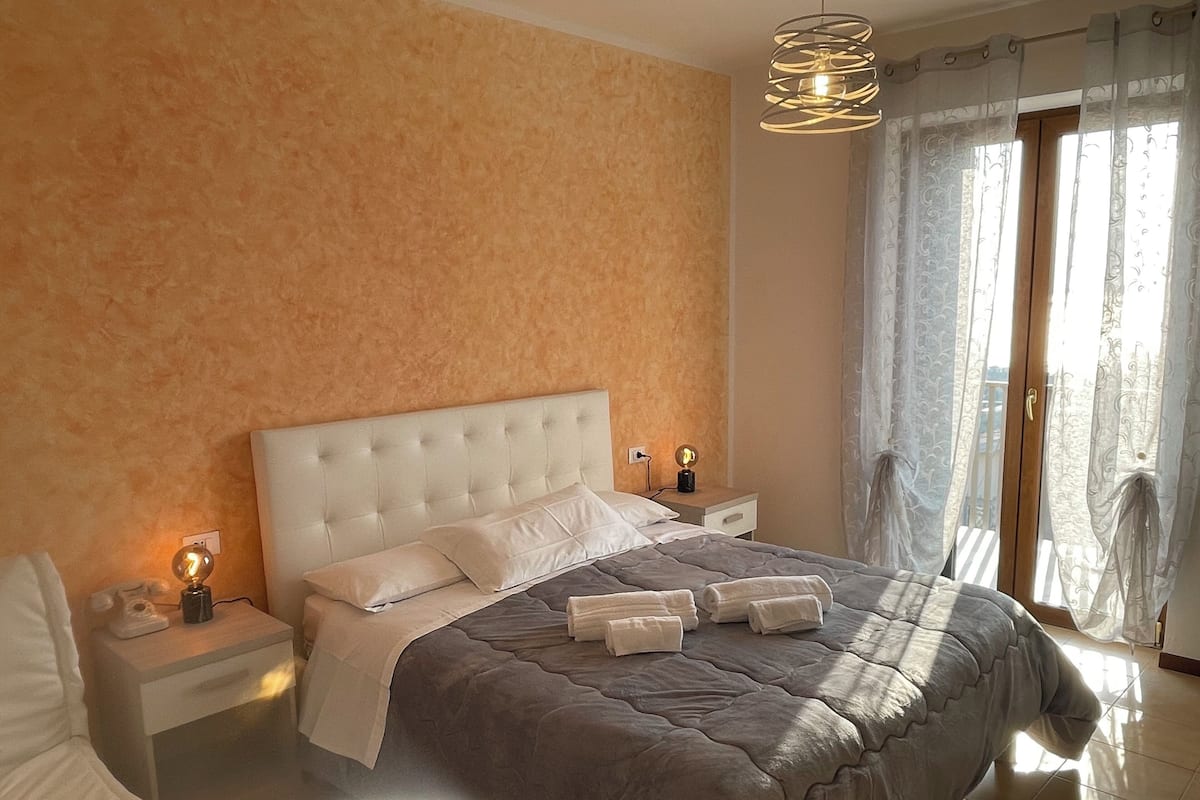Un dormitorio con una cama grande, dos mesitas de noche con lámparas, una ventana con cortinas y vistas al exterior.