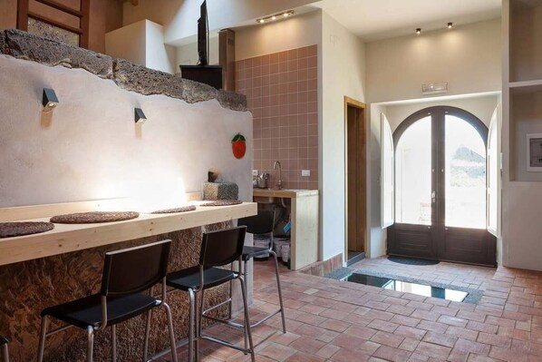 A kitchen with a stone counter, bar stools, and a view of the outside through a large arched doorway.