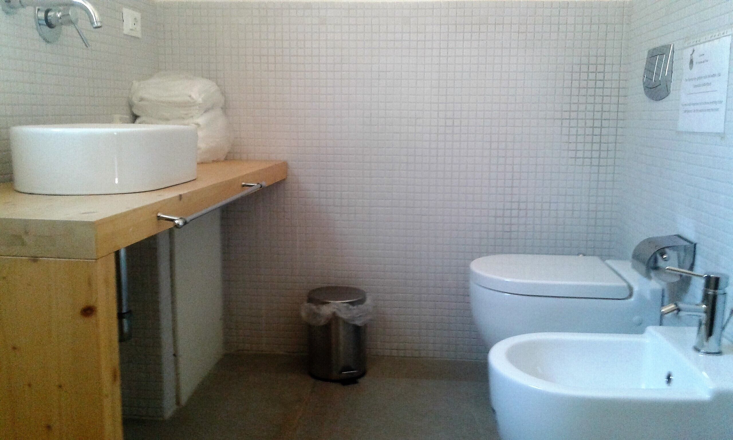 A small bathroom with a sink, toilet, and bidet. There is a trash can and a towel dispenser on the wall.