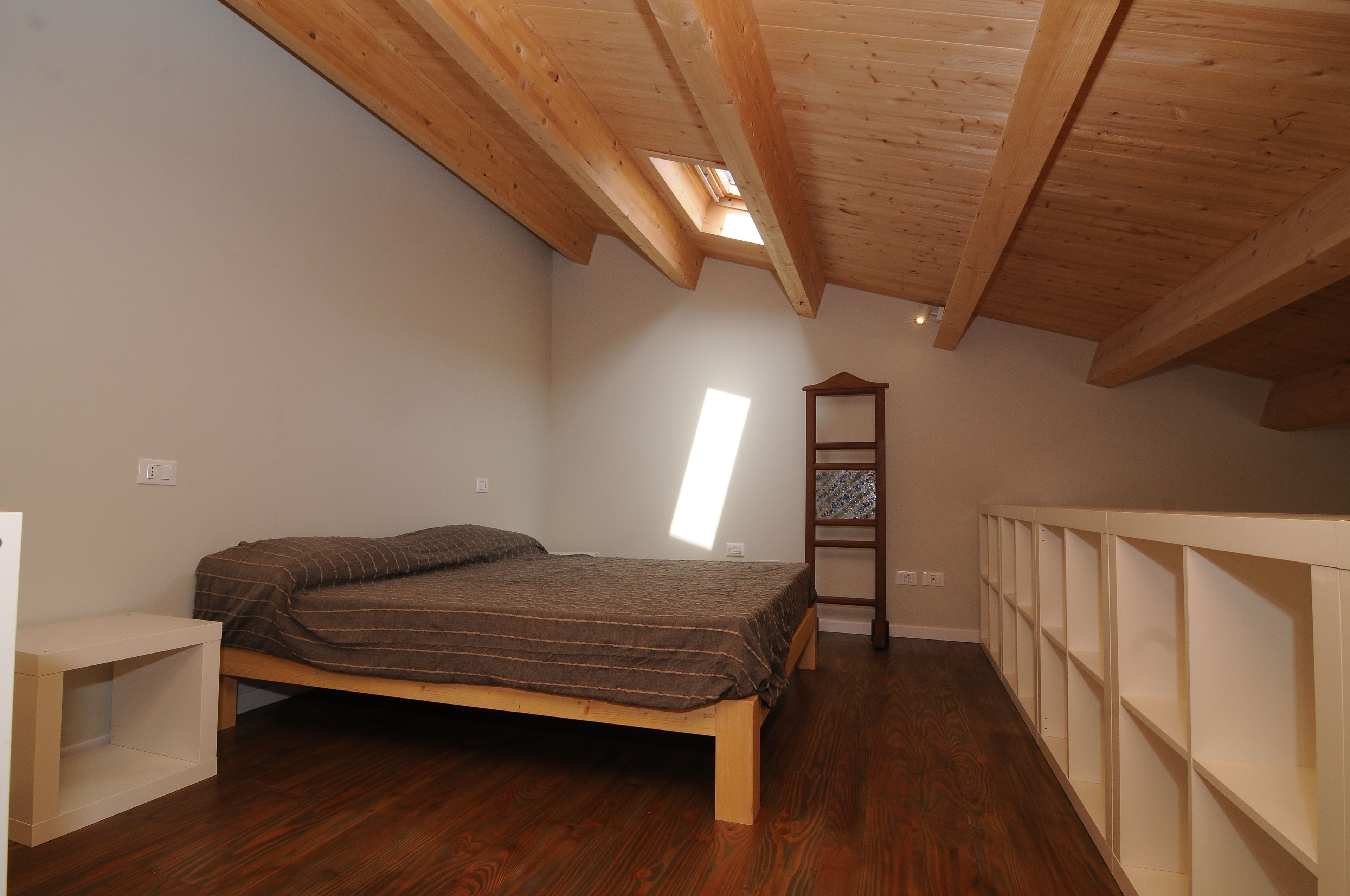 Une chambre avec un plafond en bois, une lucarne, un lit, une table de chevet et des étagères intégrées.