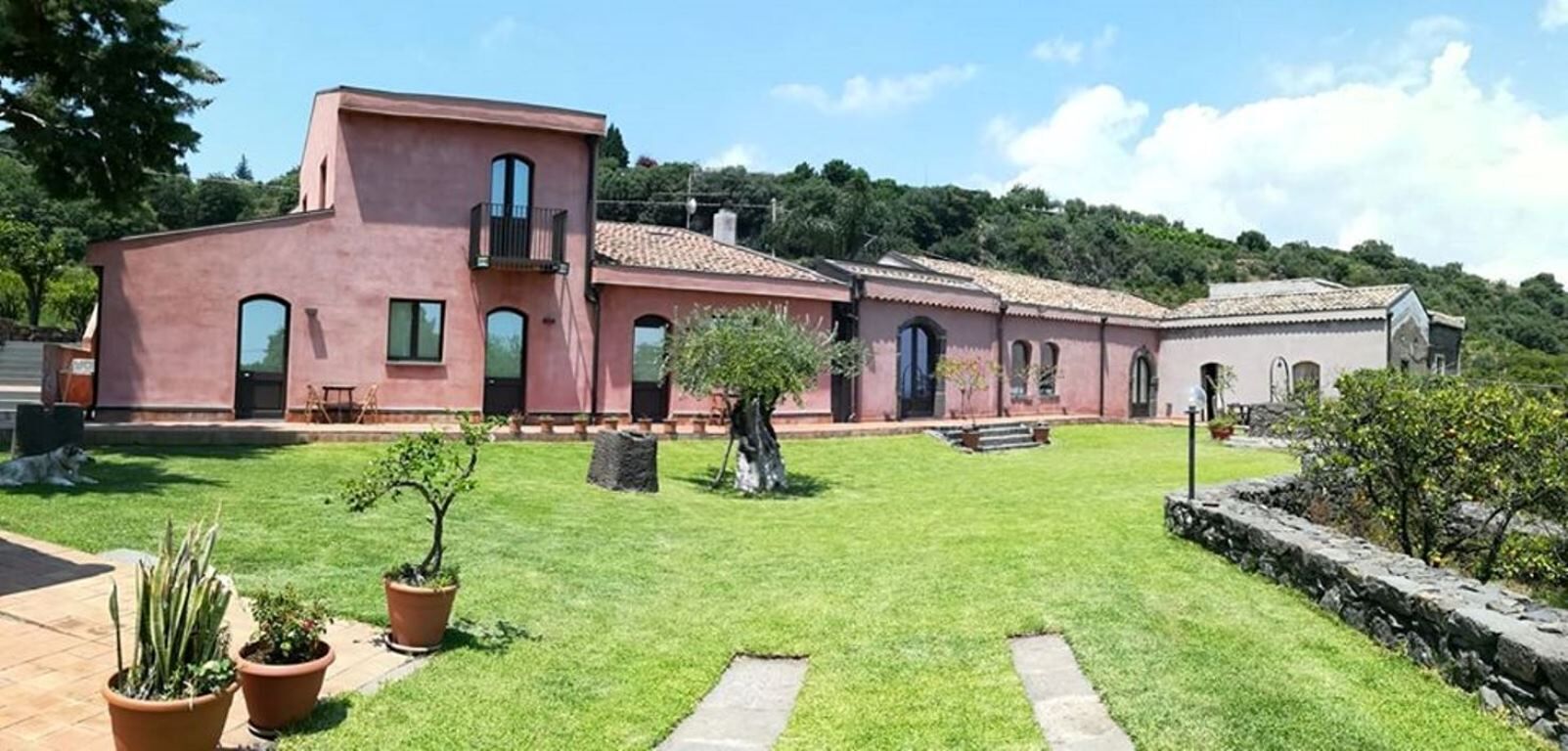A pink building with a tiled roof, a small balcony, and a well-maintained lawn with potted plants.