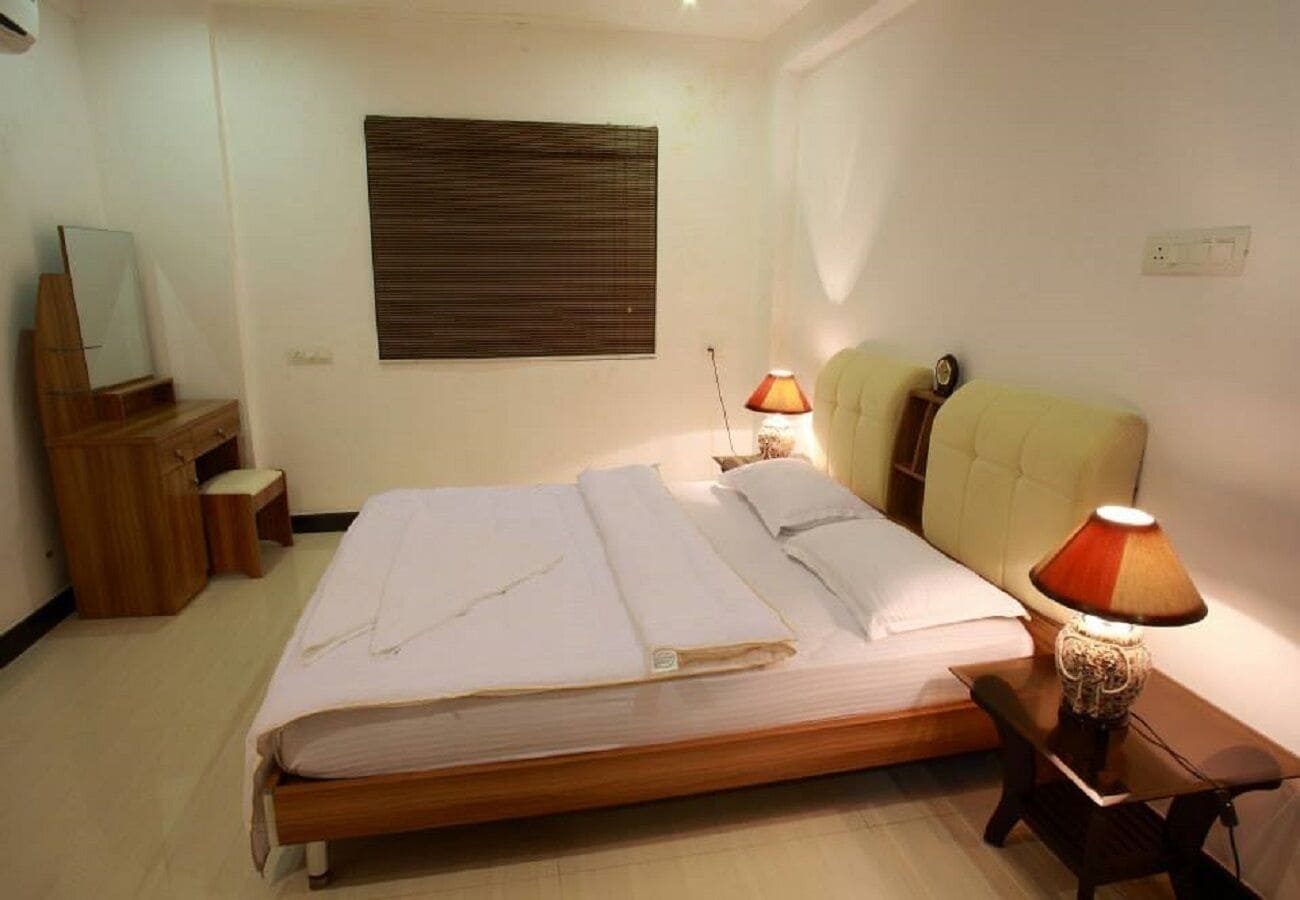 Una cama bien hecha con cabecera de madera, una mesita de noche con lámpara, un pequeño aparador de madera y un espejo a la izquierda.