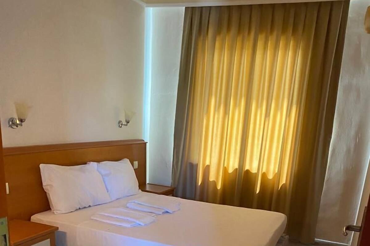 A hotel room with a bed, wooden headboard, a nightstand, and a window with curtains.