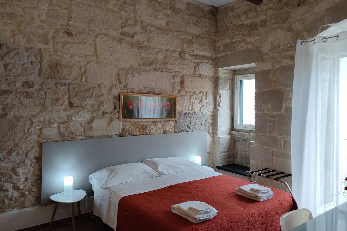 Ein Schlafzimmer mit Steinwand, einem Bett mit weißer Bettwäsche, einer roten Decke und einem Blumenbild.