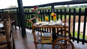 A wooden table with a vintage cart, a vase with flowers, and a tray with breakfast items on a terrace with a wooden railing and a view of greenery. - Treetops (Aberdare National Park)