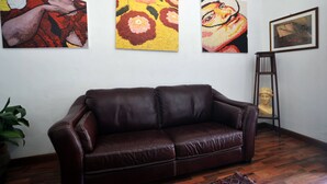 Een kamer met een bruine leren bank, drie kleurrijke schilderijen aan de muur en een buste op een voetstuk.