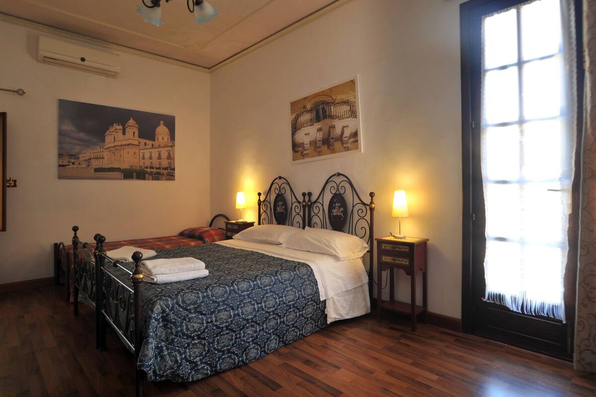 Une chambre à coucher avec un lit, deux tables de chevet avec des lampes, une fenêtre avec des rideaux et deux tableaux accrochés au mur.