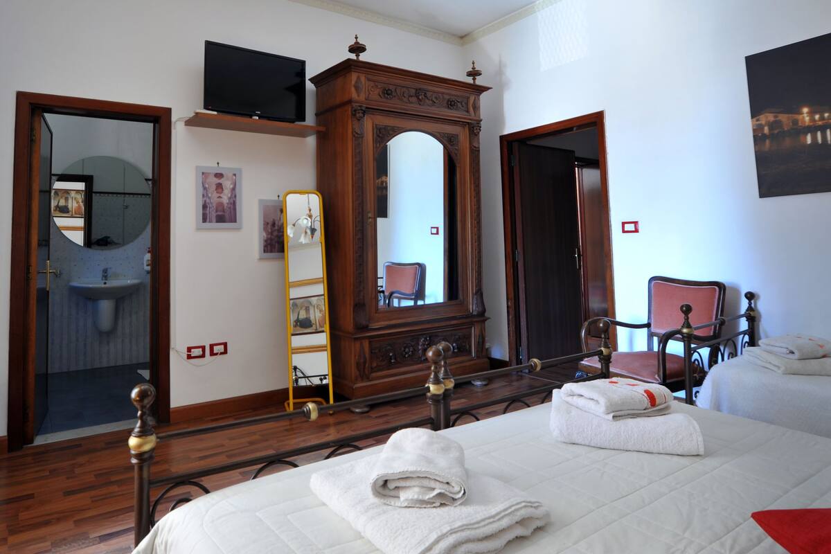 Une chambre d’hôtel avec un lit, une armoire en bois, un miroir, une télévision et une salle de bain visible à travers une porte ouverte.