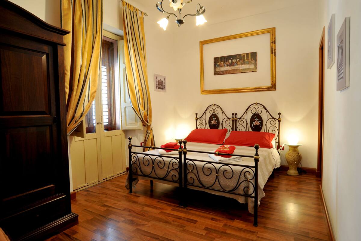 Une chambre avec un plancher en bois, un cadre de lit en métal, une table de chevet avec une lampe, une fenêtre avec des rideaux et un tableau encadré au mur.