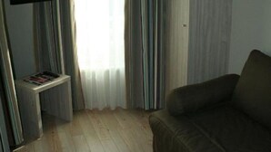 A room with a wooden floor, a brown sofa, a TV on a stand, and a window with sheer curtains. - Hôtel L'Antinéa (Saint-Malo)