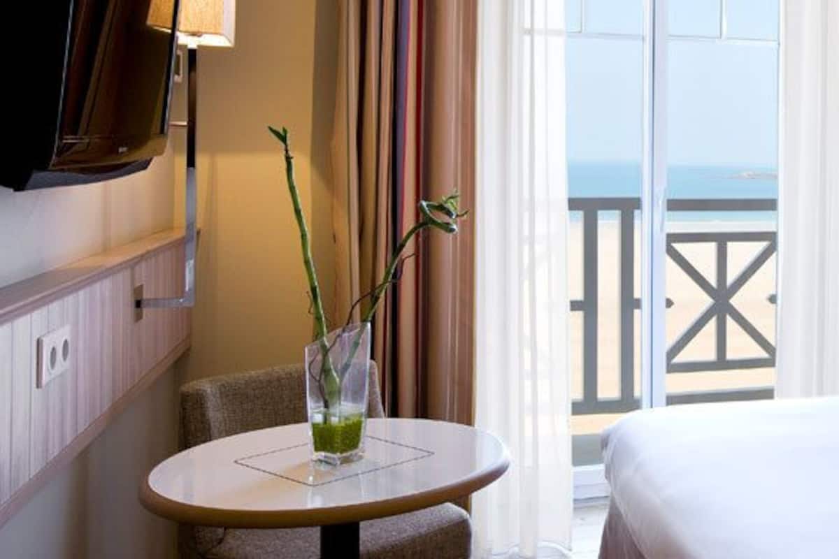 A hotel room with a bed, a table with a vase, a TV, and a balcony with a railing.