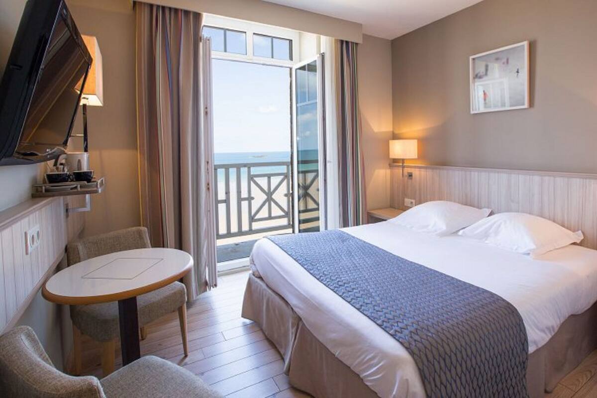 A hotel room with a bed, a small table, a chair, a TV, a balcony with a railing, and a painting on the wall.