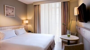 A hotel room with a large bed, a bedside table with a lamp, a small round table with a vase, and a television mounted on the wall. - Hôtel L'Antinéa (Saint-Malo)