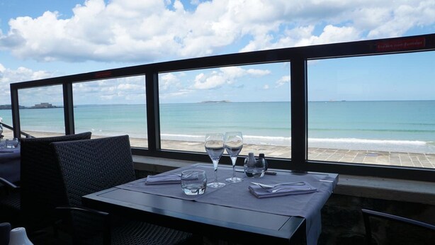 A table with two wine glasses and a water glass, set against a backdrop of a beach and ocean view. - Hôtel L'Antinéa (Saint-Malo)