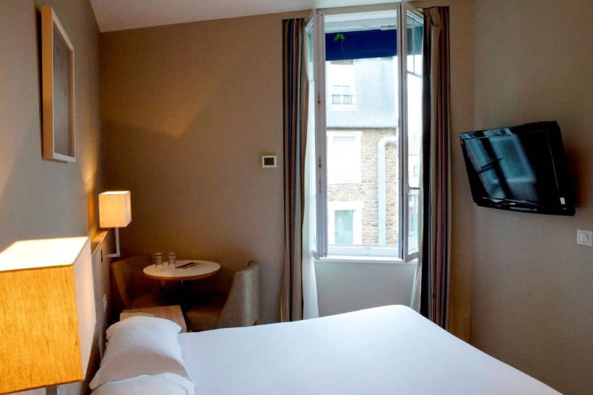 A hotel room with a bed, a TV mounted on the wall, a small table with a lamp, and a window with curtains.