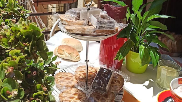 A table with a variety of pastries and bread, a potted plant, and a glass of drink, set against a building with stone walls and wooden shutters. - B&B Le Quattro Stagioni (Agrigento)
