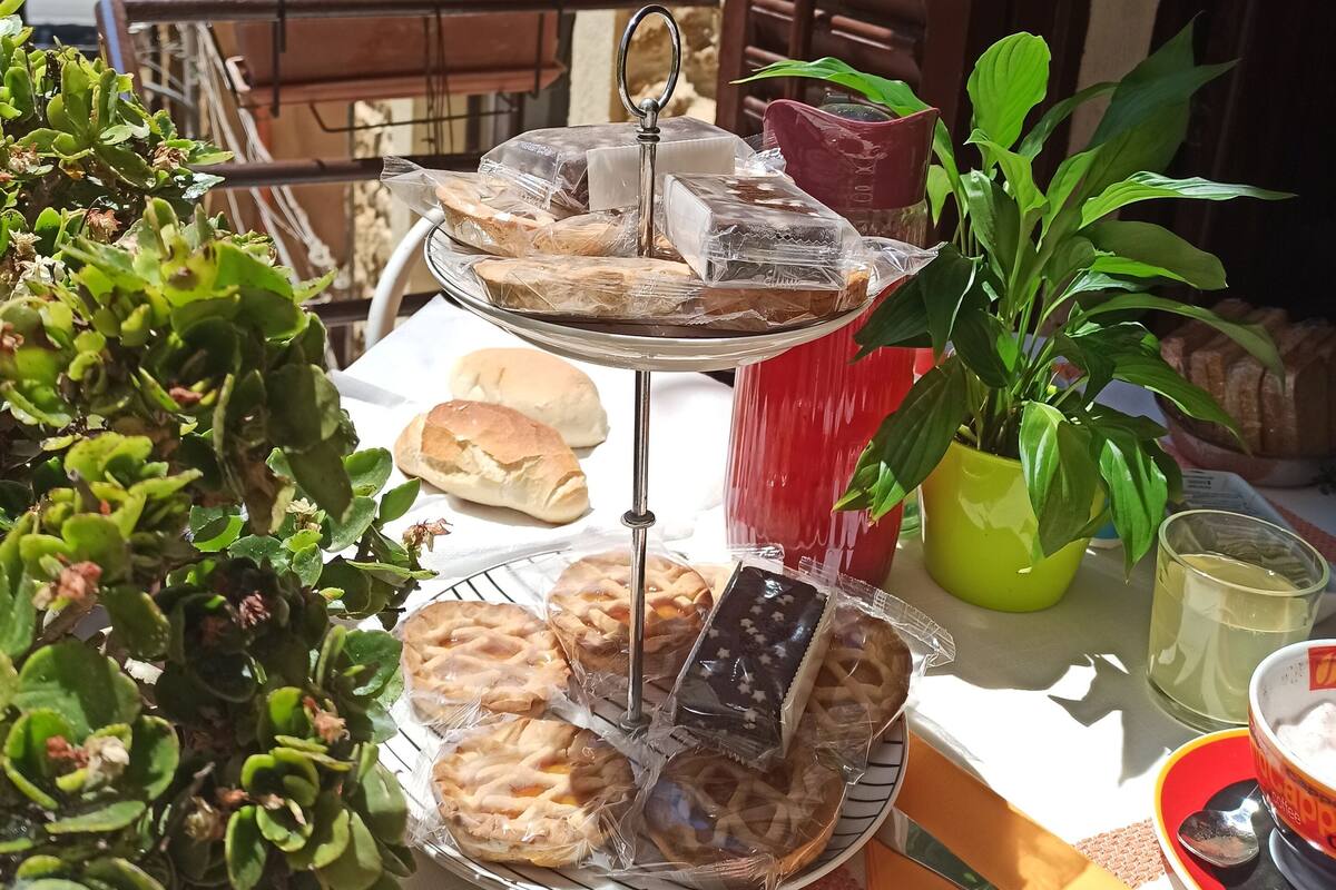 Uma mesa com diversos pães e doces, uma planta em vaso, e um copo de bebida, tudo em frente a um edifício com paredes de pedra e persianas de madeira.