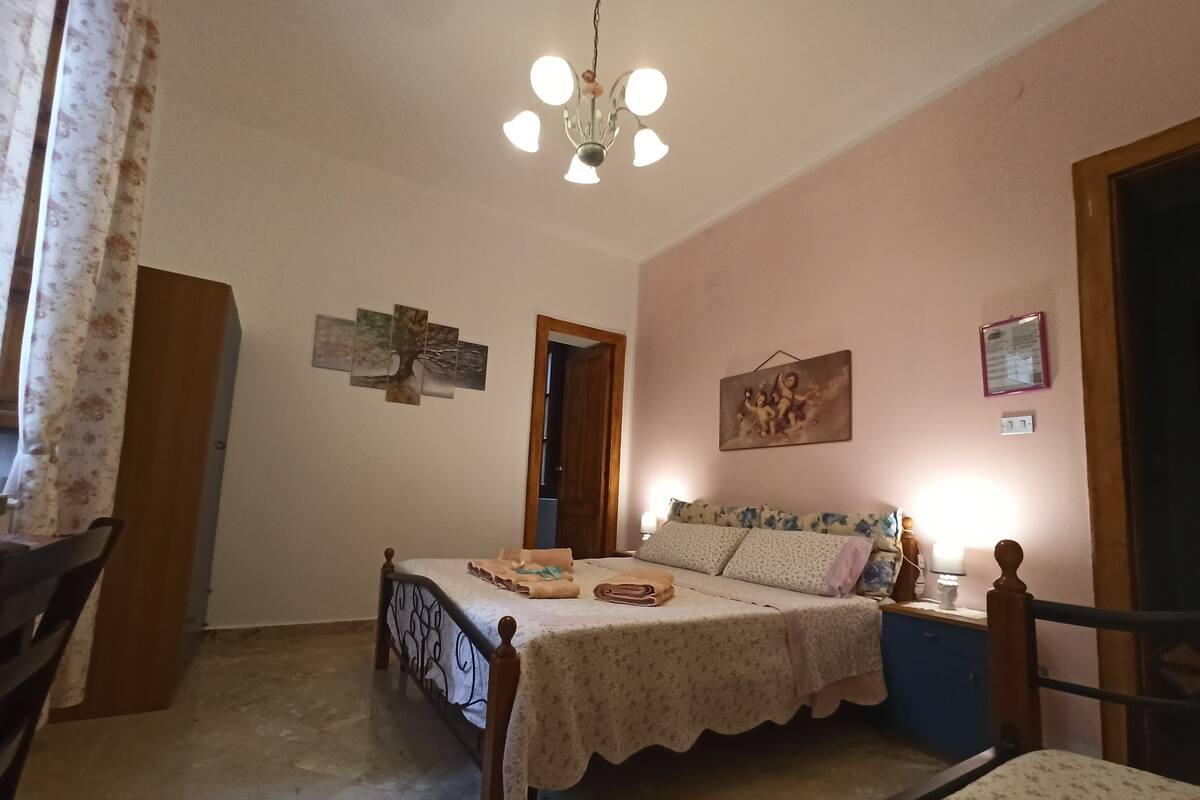 Quarto com cama, mesa de cabeceira, cadeira, luminária no teto e porta.