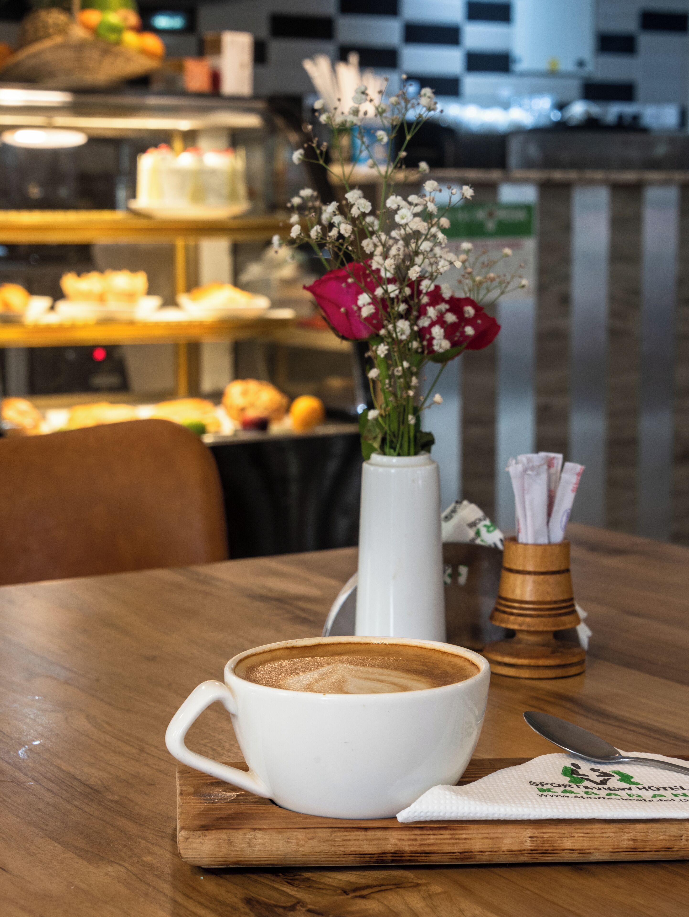 Una taza blanca de café sobre una mesa de madera con un jarrón blanco con flores y una cuchara.