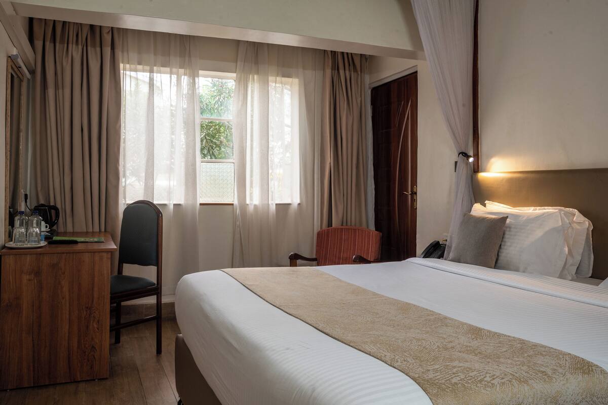 A hotel room with a large bed, a desk with a chair, a window with curtains, and a bedside lamp.