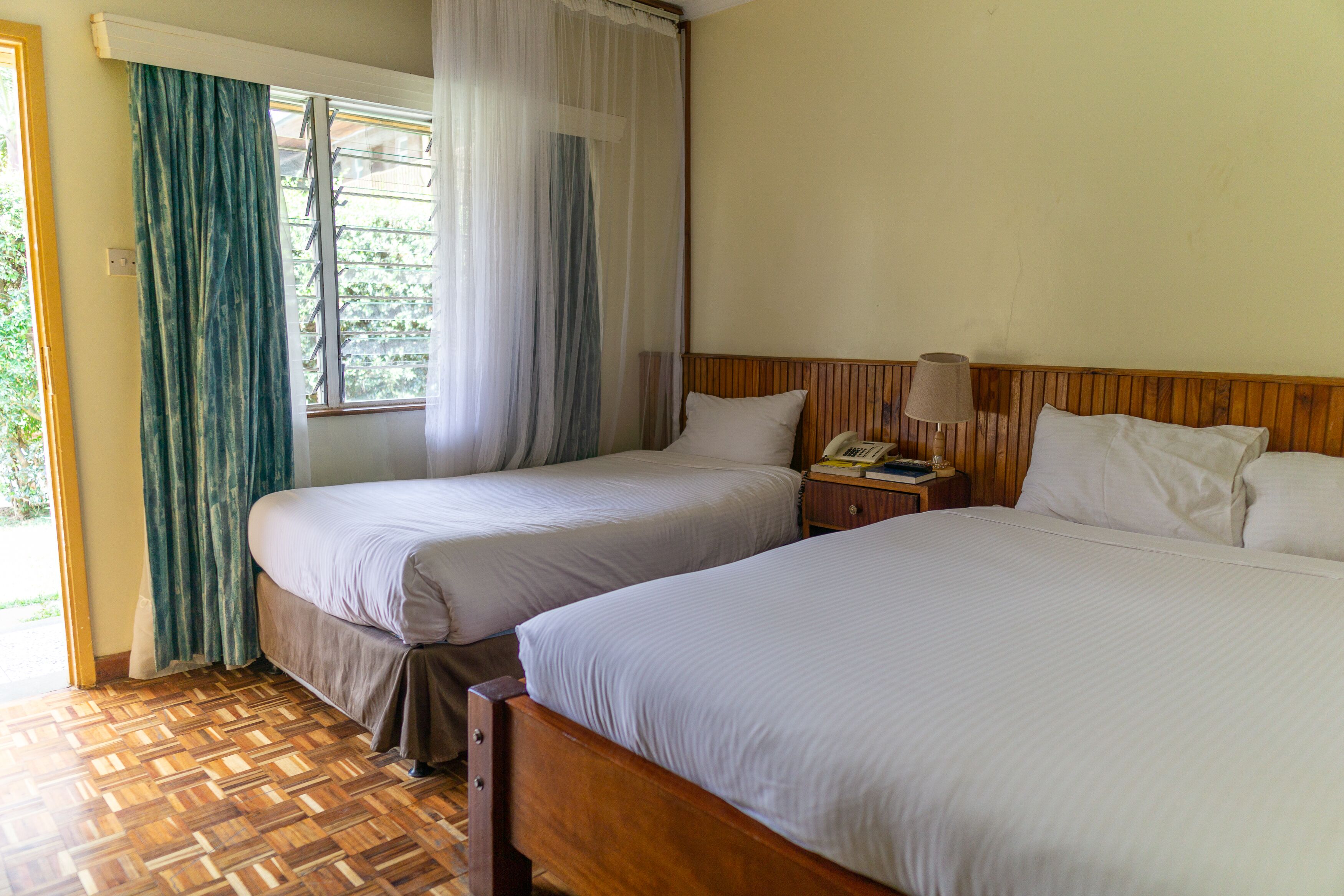 Une chambre d’hôtel avec deux lits, une tête de lit en bois, une table de chevet avec une lampe et une fenêtre avec des rideaux légers.