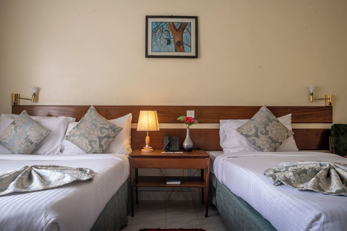 A hotel room with two beds, a nightstand with a lamp, a vase with flowers, and a framed picture on the wall.