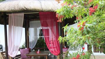 A covered outdoor seating area with a table and chairs, surrounded by potted plants and a thatched roof structure.