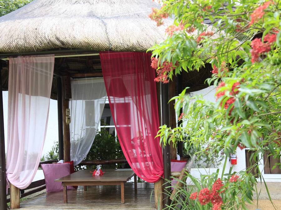 A covered outdoor seating area with a table and chairs, surrounded by potted plants and a thatched roof structure.