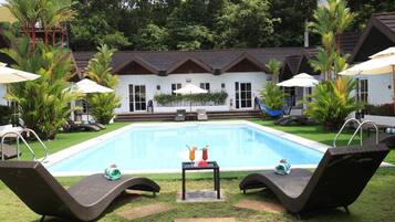 Une piscine entourée de chaises longues et de parasols, elle-même bordée d’une pelouse bien entretenue et d’un bâtiment dont le toit est recouvert de tuiles.