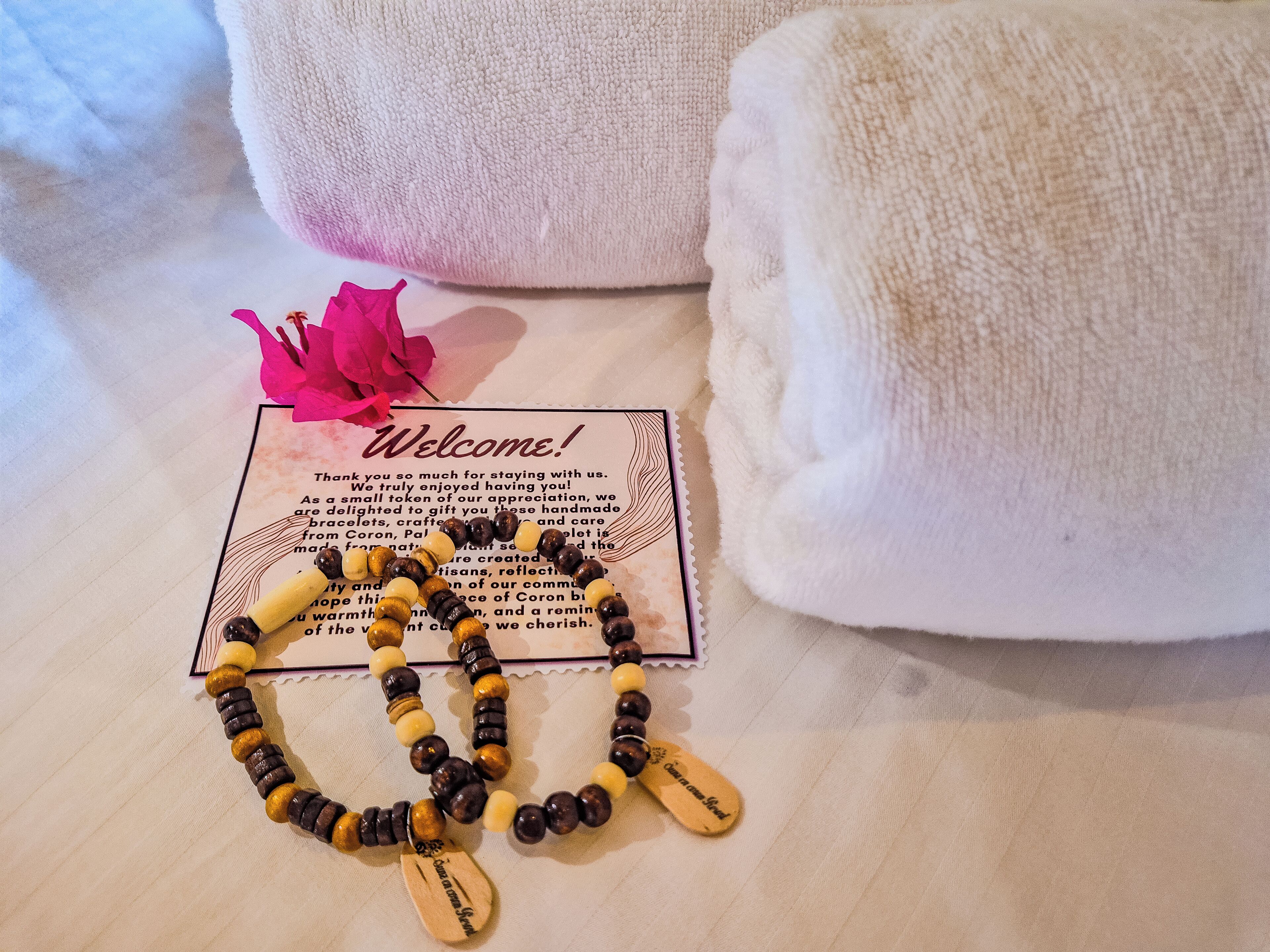 A welcome card, a beaded bracelet, and a pink flower on a bed with white towels.