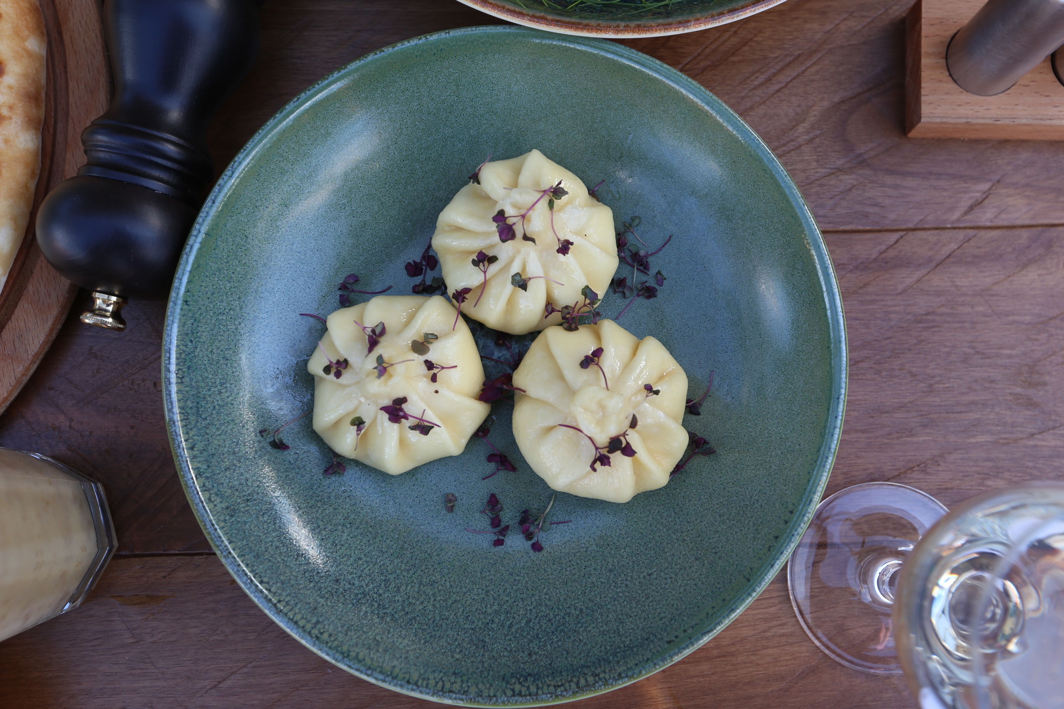 Un plato de dumplings decorado con hierbas, un molinillo de pimienta y un vaso de agua sobre una mesa de madera.