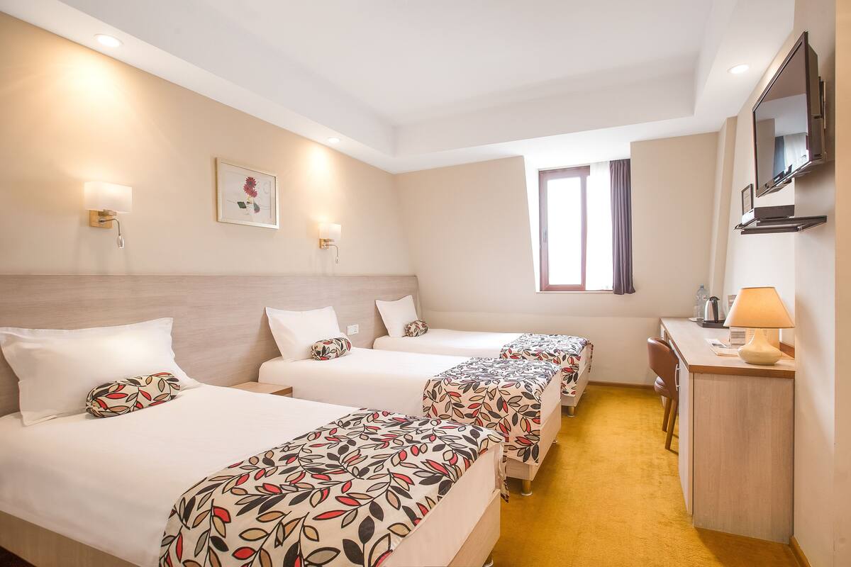 A hotel room with three beds, a desk, a chair, a TV, and a window.