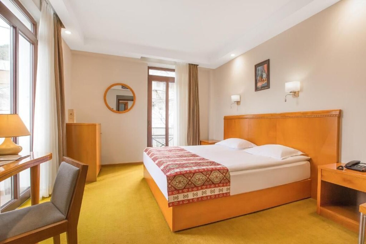 A hotel room with a bed, a desk, a chair, a mirror, and a wall-mounted light.