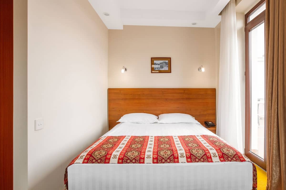A hotel room with a wooden headboard, a bed with a patterned bedspread, two wall-mounted lights, a framed picture on the wall, and a window with curtains.