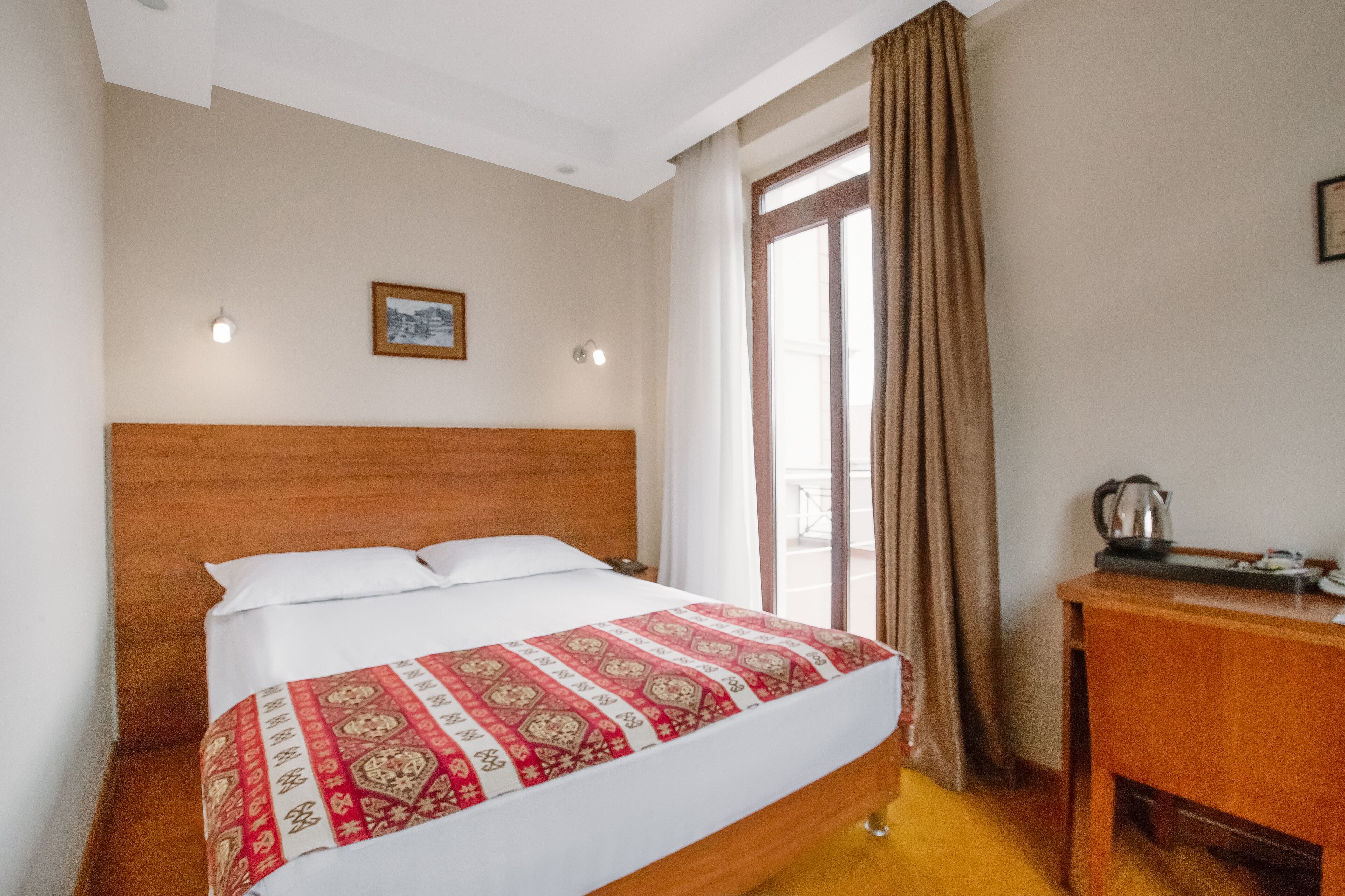 Habitació d'hotel amb un llit de fusta, una petita taula de nit amb un búl i una finestra amb cortines.