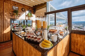 Un bufé de desayuno con diversos alimentos y vistas a la ciudad a través de amplias ventanas.