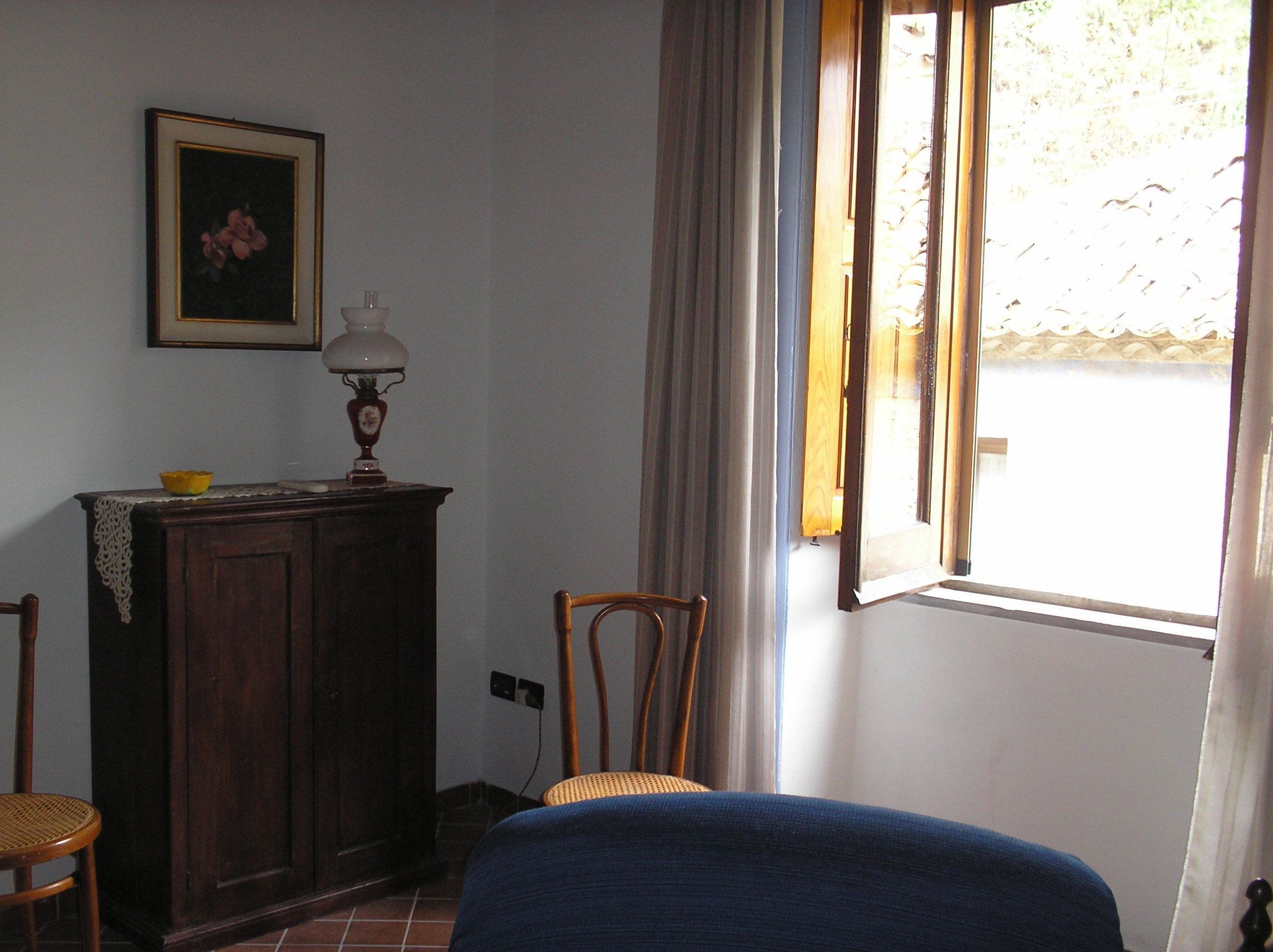 Una habitación con un armario de madera, una silla, una cama, una ventana con cortinas y un cuadro en la pared.