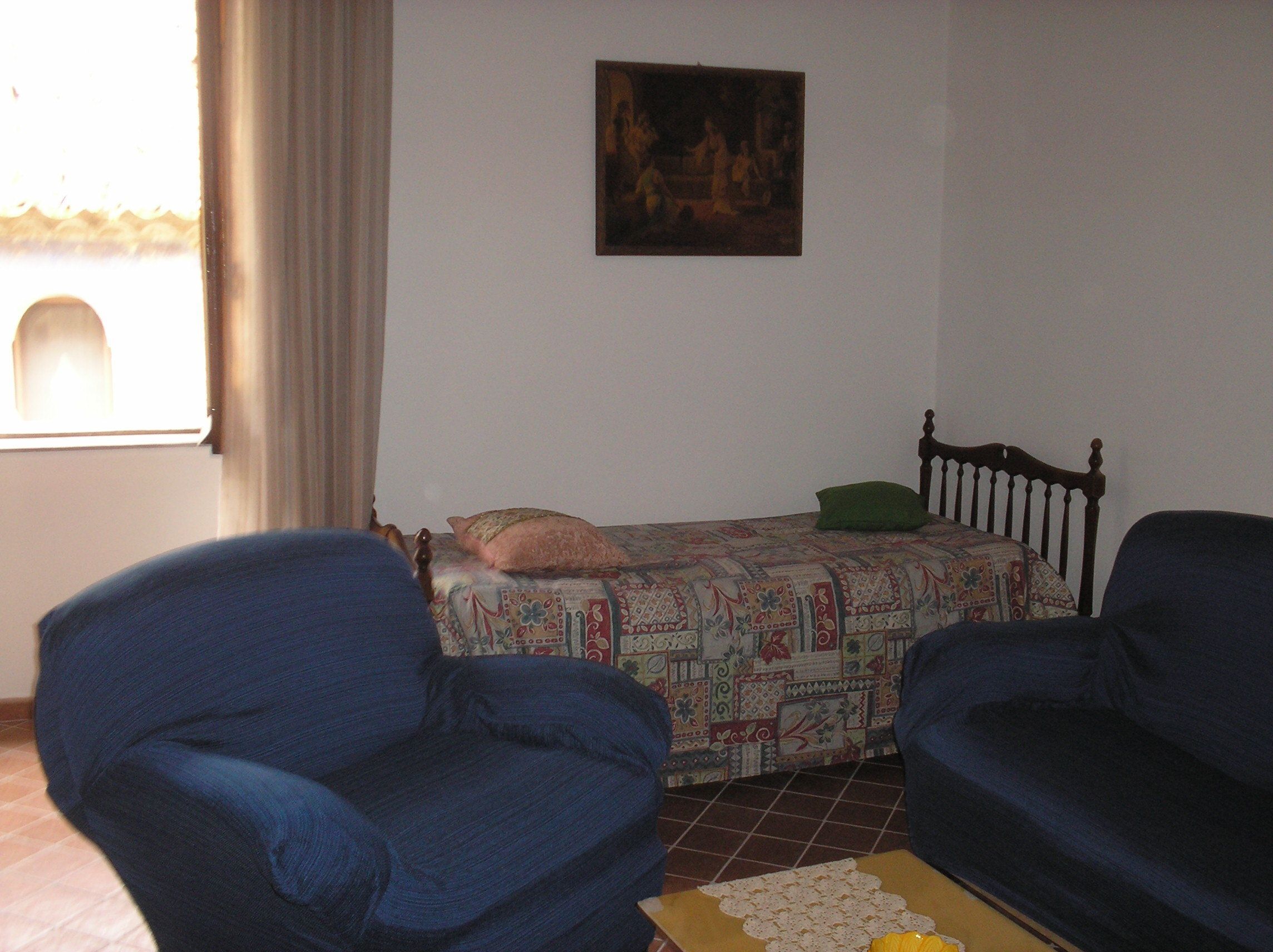 A room with a bed, two blue armchairs, a small table with a doily, and a painting on the wall.
