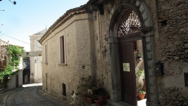 Une ruelle étroite pavée, bordée d’un bâtiment historique remarquable par son arc orné et ses plantes en pot.