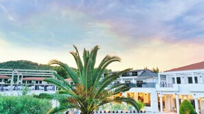 A hotel with a modern architectural style, featuring a prominent palm tree in the foreground, a balcony with outdoor seating, and a view of a hilly landscape in the background.